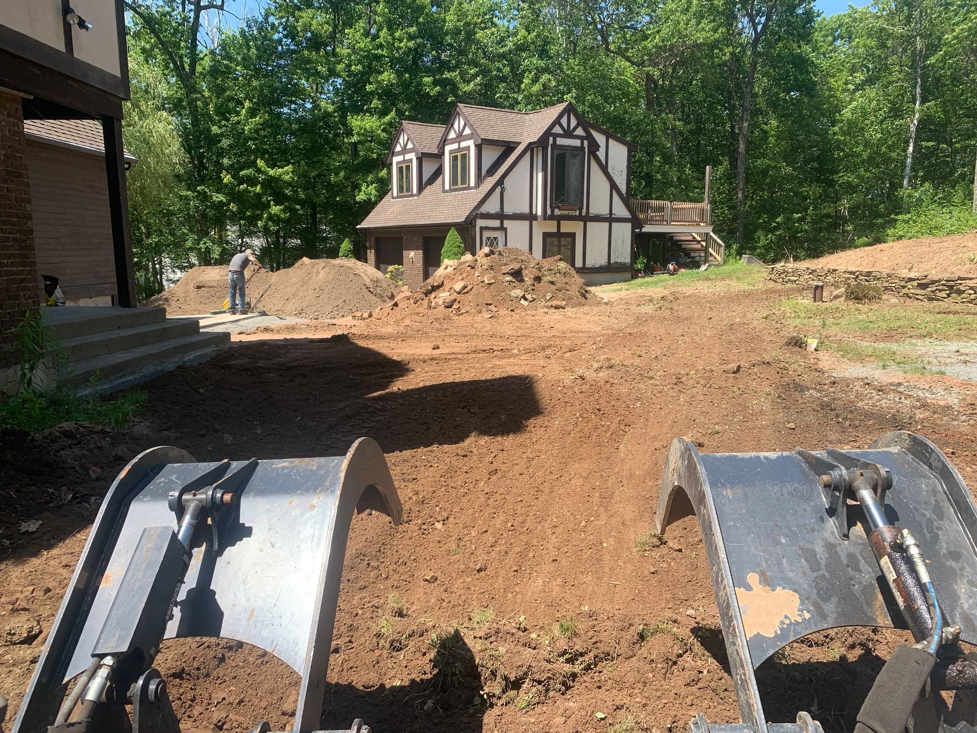 A backhoe digging in a cleared dirt yard near a Tudor-style house and a person with a shovel.