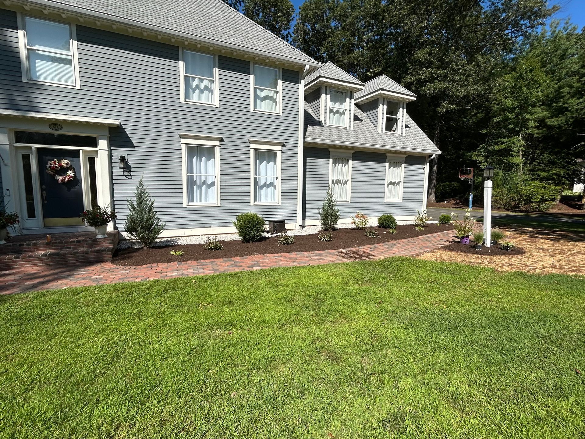Two-story gray house with green lawn, brick walkway, and landscaped garden beds.