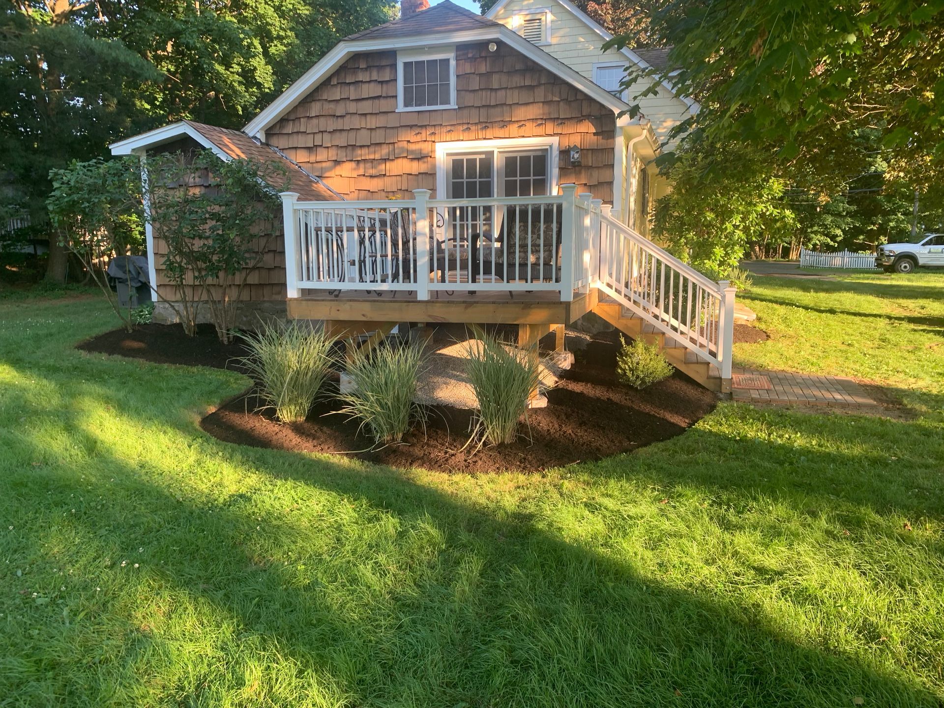 Cottage with a white deck, surrounded by fresh mulch and shrubs, set in a grassy yard.
