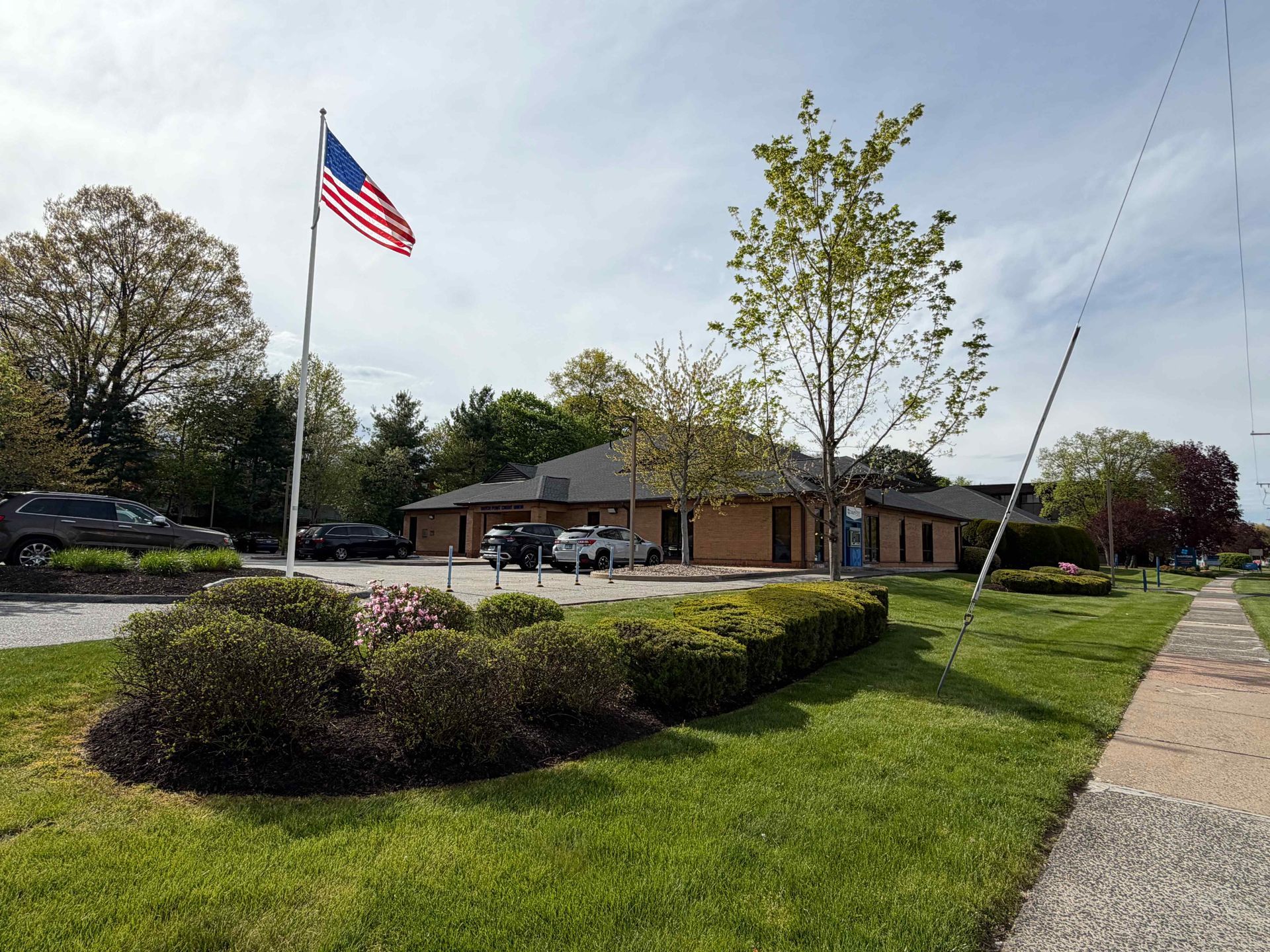 Small brick building beside a grassy lawn, with a large American flag on a pole and a sidewalk in front.
