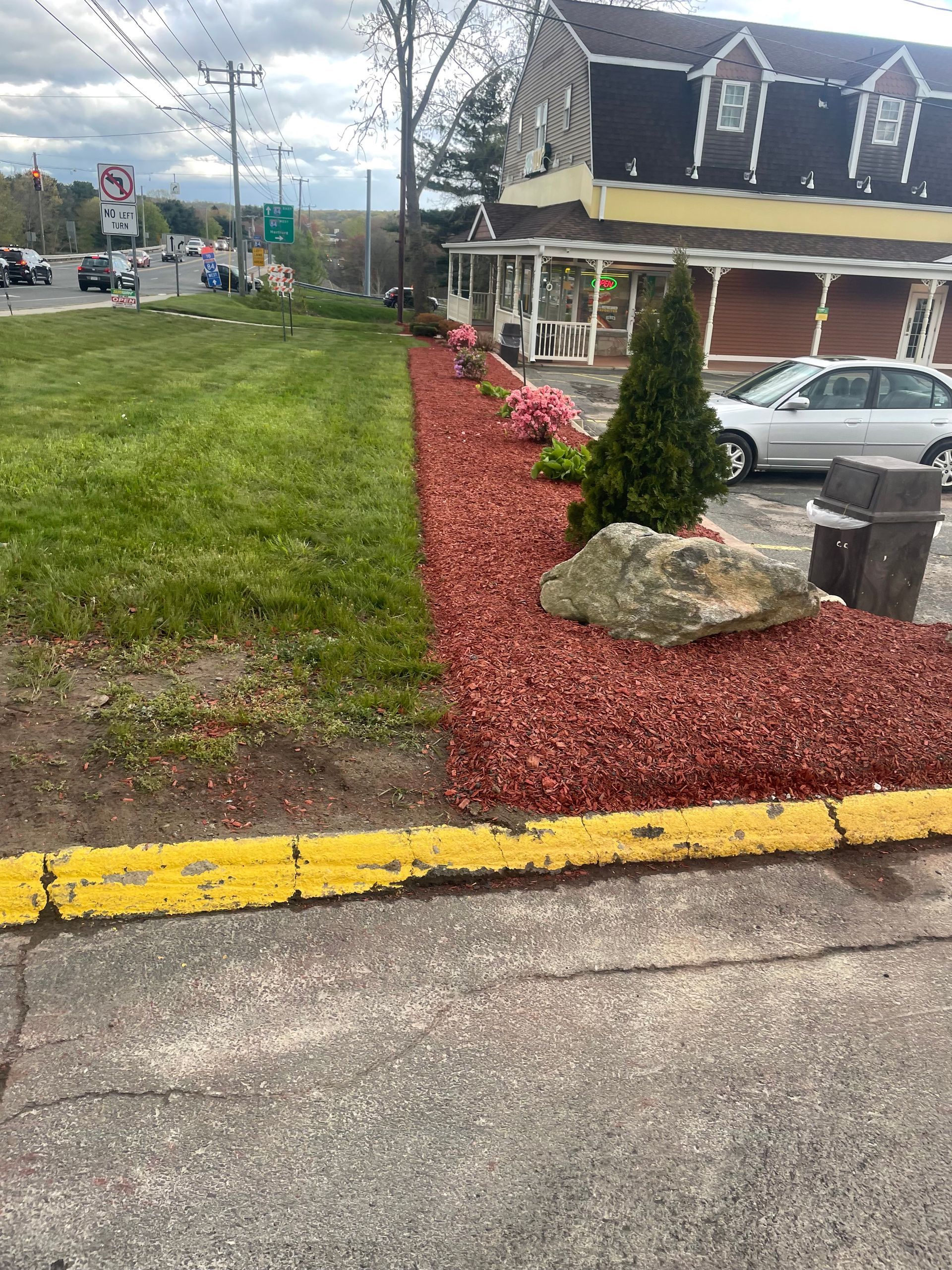 A landscaped bed with red mulch, flowers, and a large rock in front of a building with cars and street beyond.