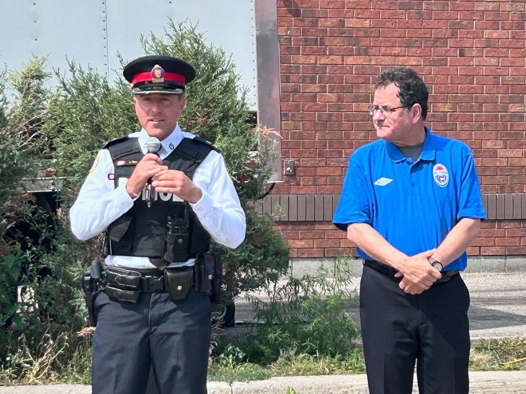 Two men are standing next to each other in front of a brick building.