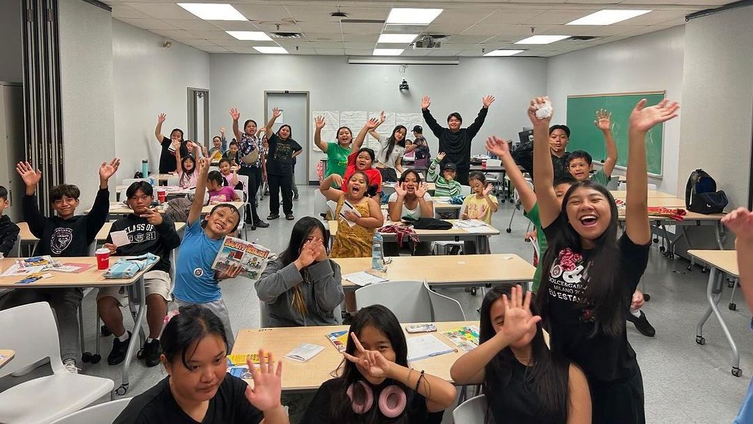 A group of children are standing in a classroom with their hands in the air.