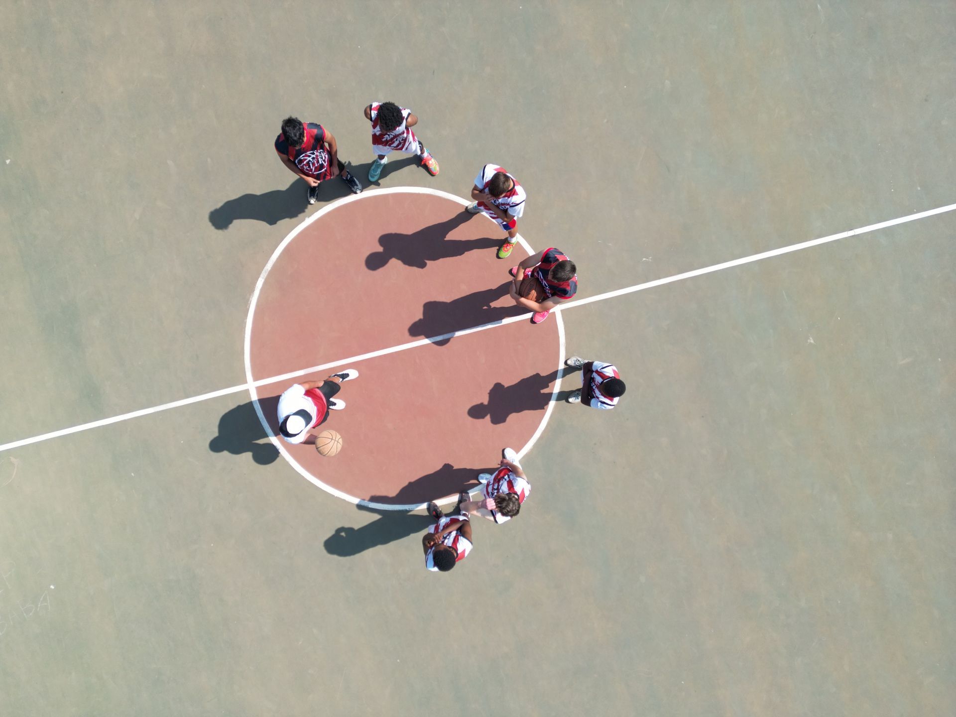 Players on basketball court, some in maroon, near center circle, shadows cast by sun.