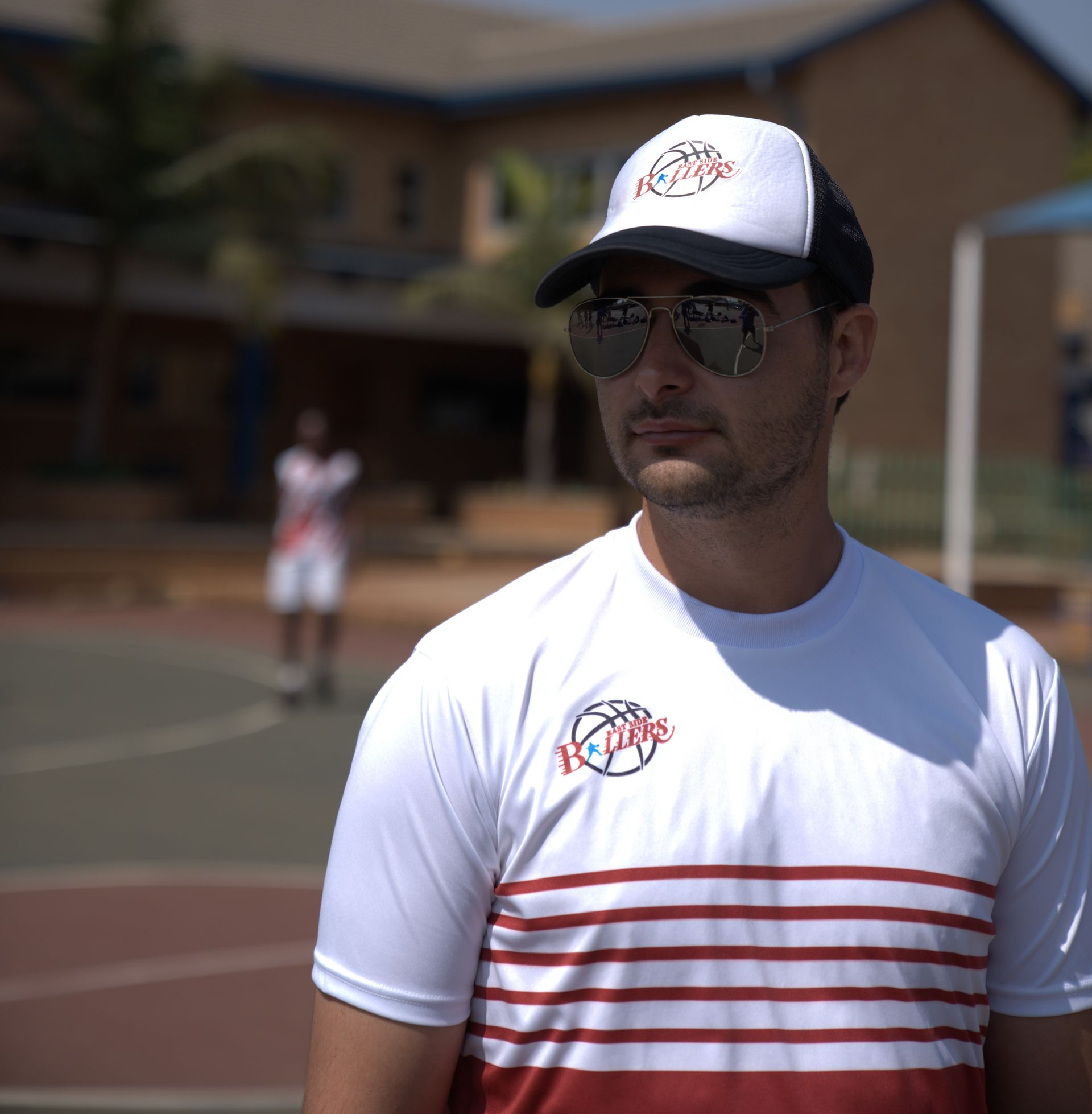 Man wearing hat and sunglasses, standing on basketball court, red and white shirt, another person in background.