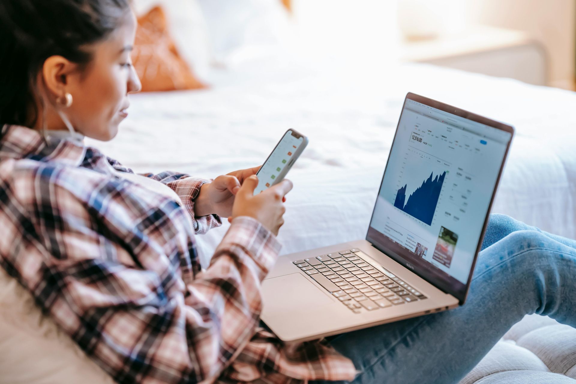 A woman is sitting on a bed using a laptop and a cell phone.