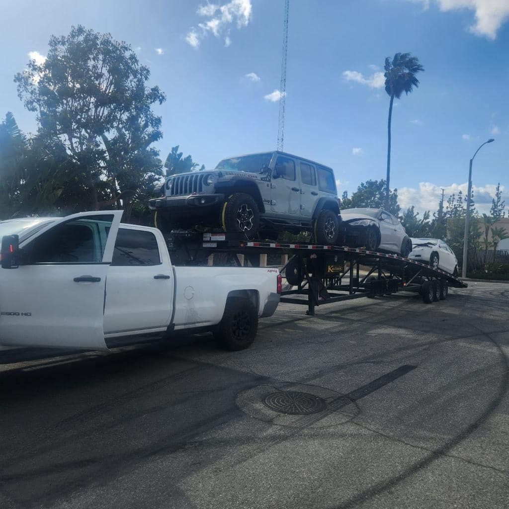 White truck hauling a trailer with a grey Jeep and two other cars on it, outdoors.