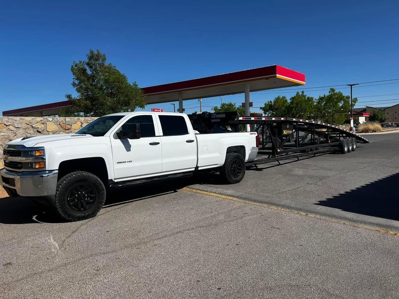 White pickup truck towing a black car trailer at a gas station under a blue sky.