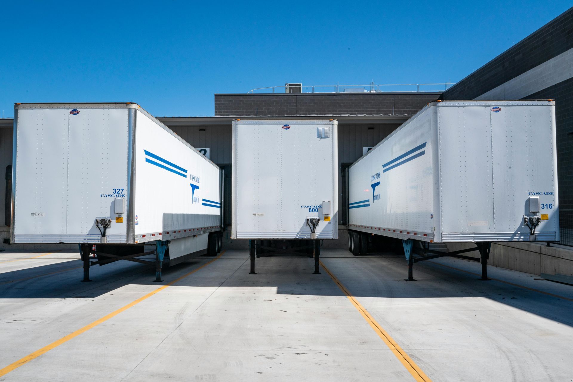 Three white semi-truck trailers parked at a loading dock against a blue sky.