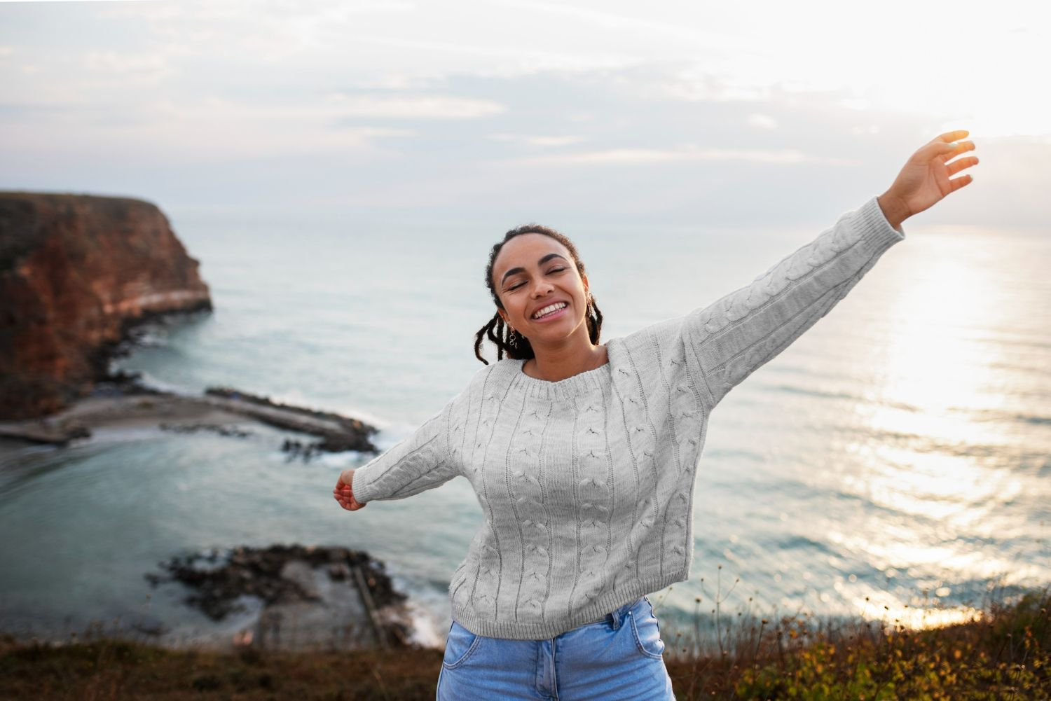 A woman is standing on top of a hill with her arms outstretched in front of the ocean.