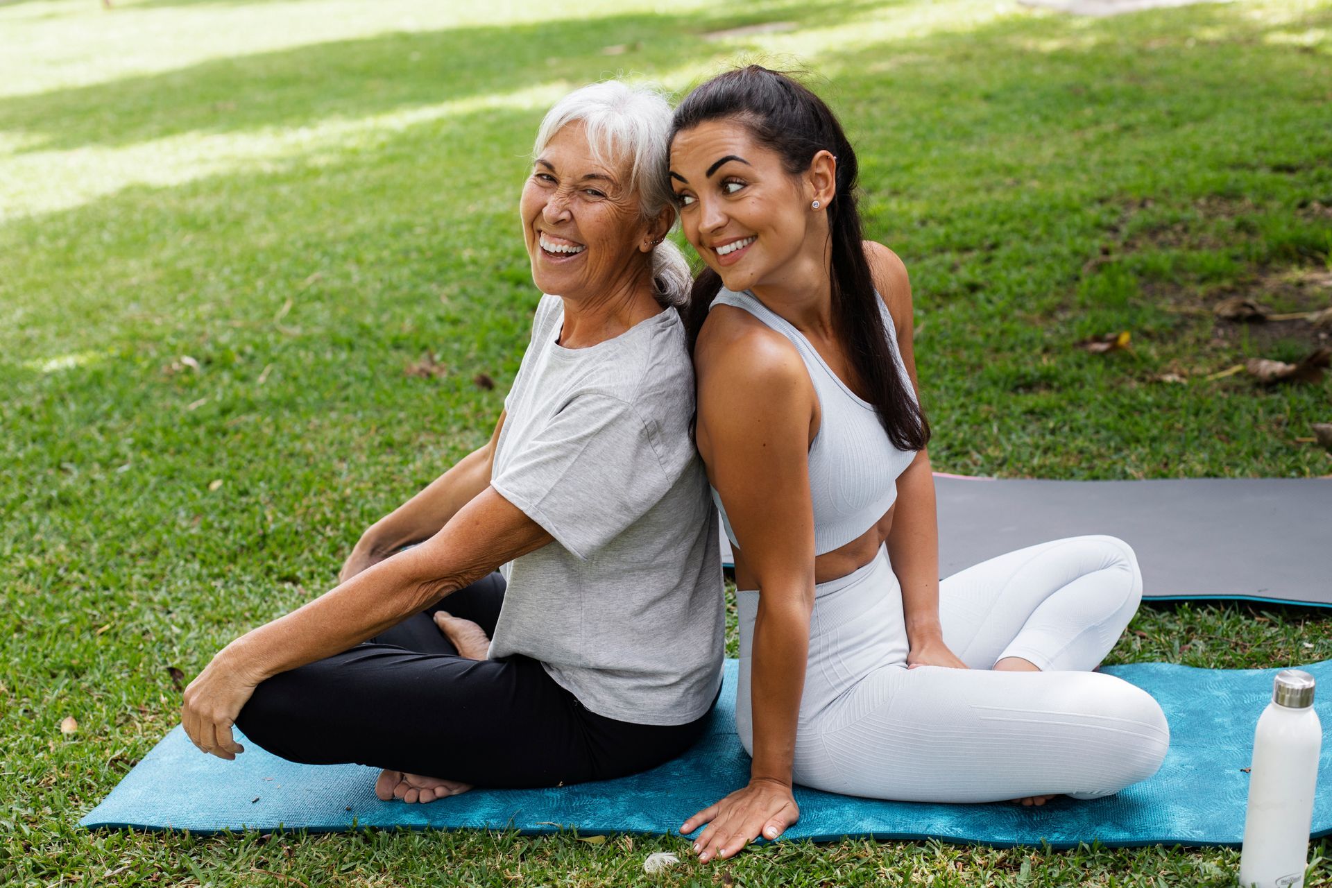 two women are sitting back to back on yoga mats in a park .