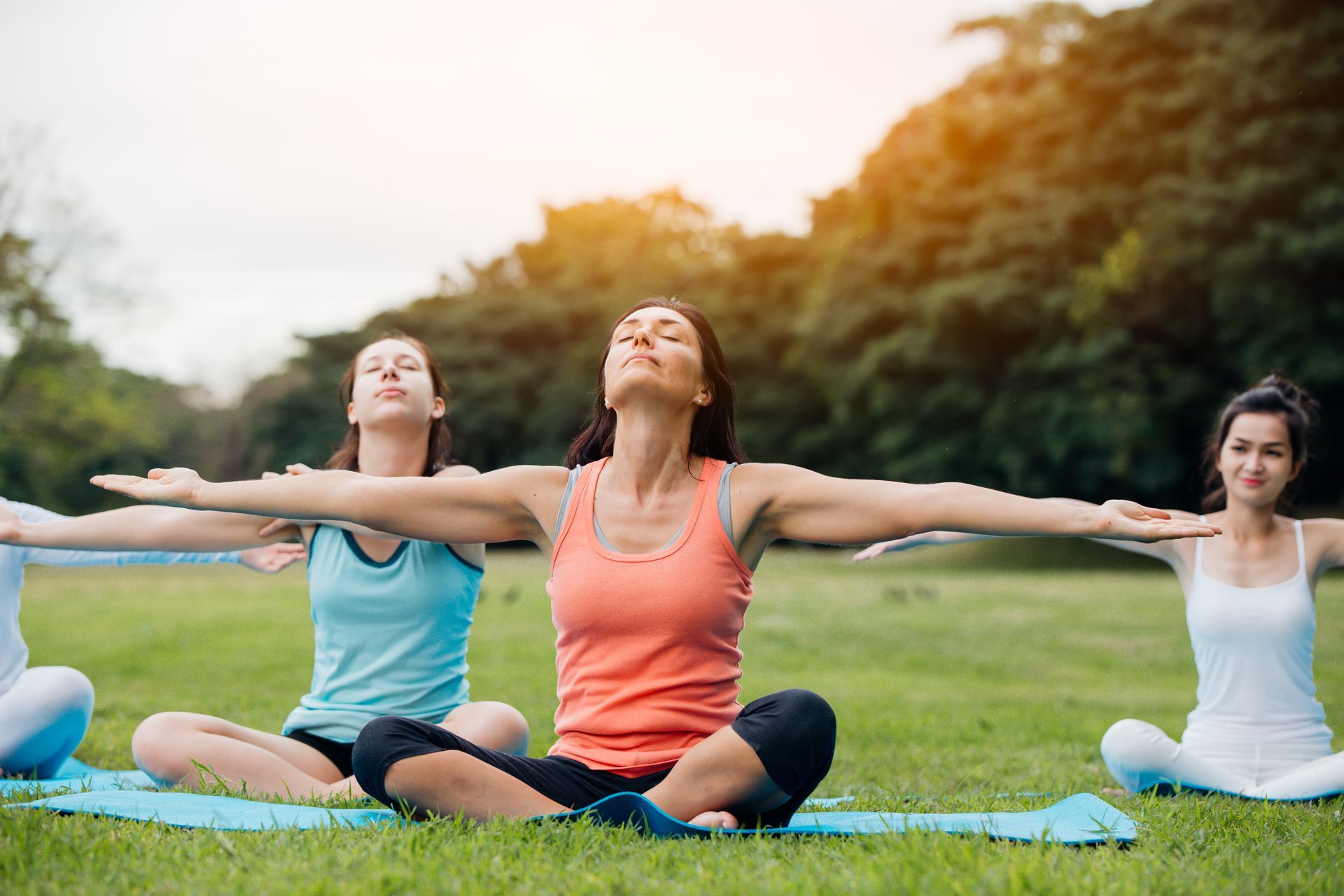 three women are sitting on yoga mats in a park with their arms outstretched .