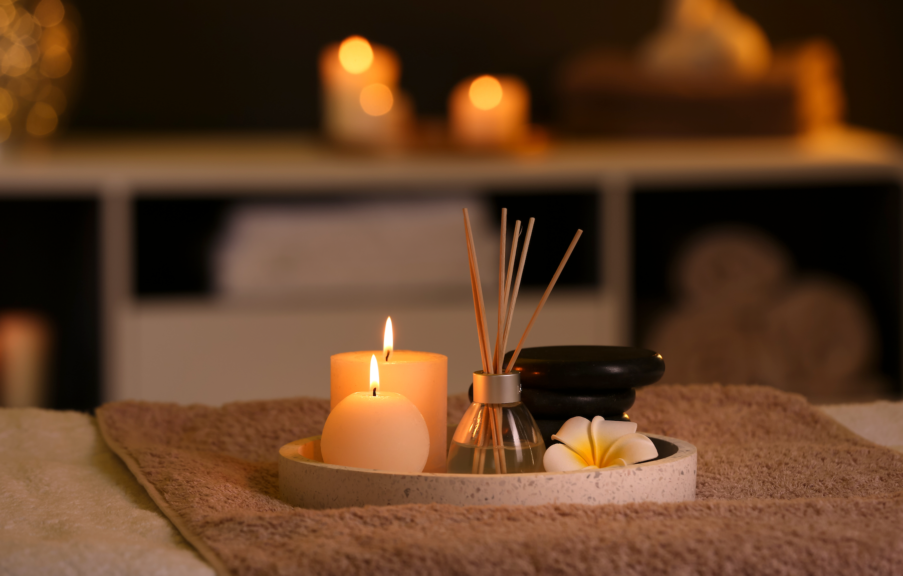 A tray with candles , reed diffusers and a flower on a towel on a bed.