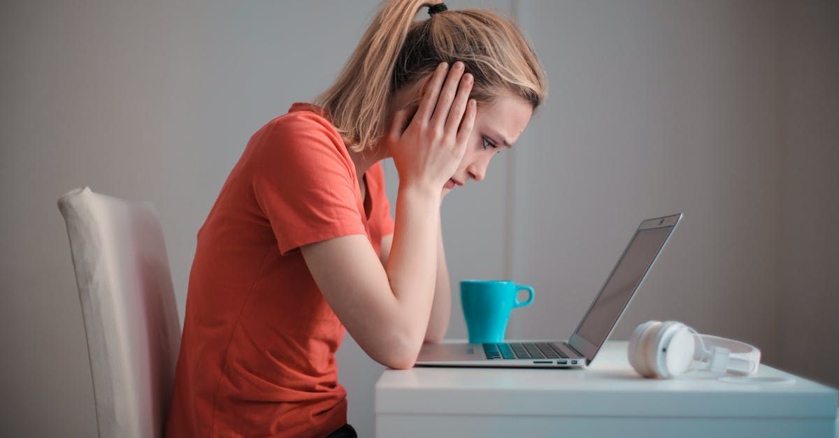 Uma mulher está sentada em uma mesa com a cabeça apoiada nas mãos em frente a um laptop.