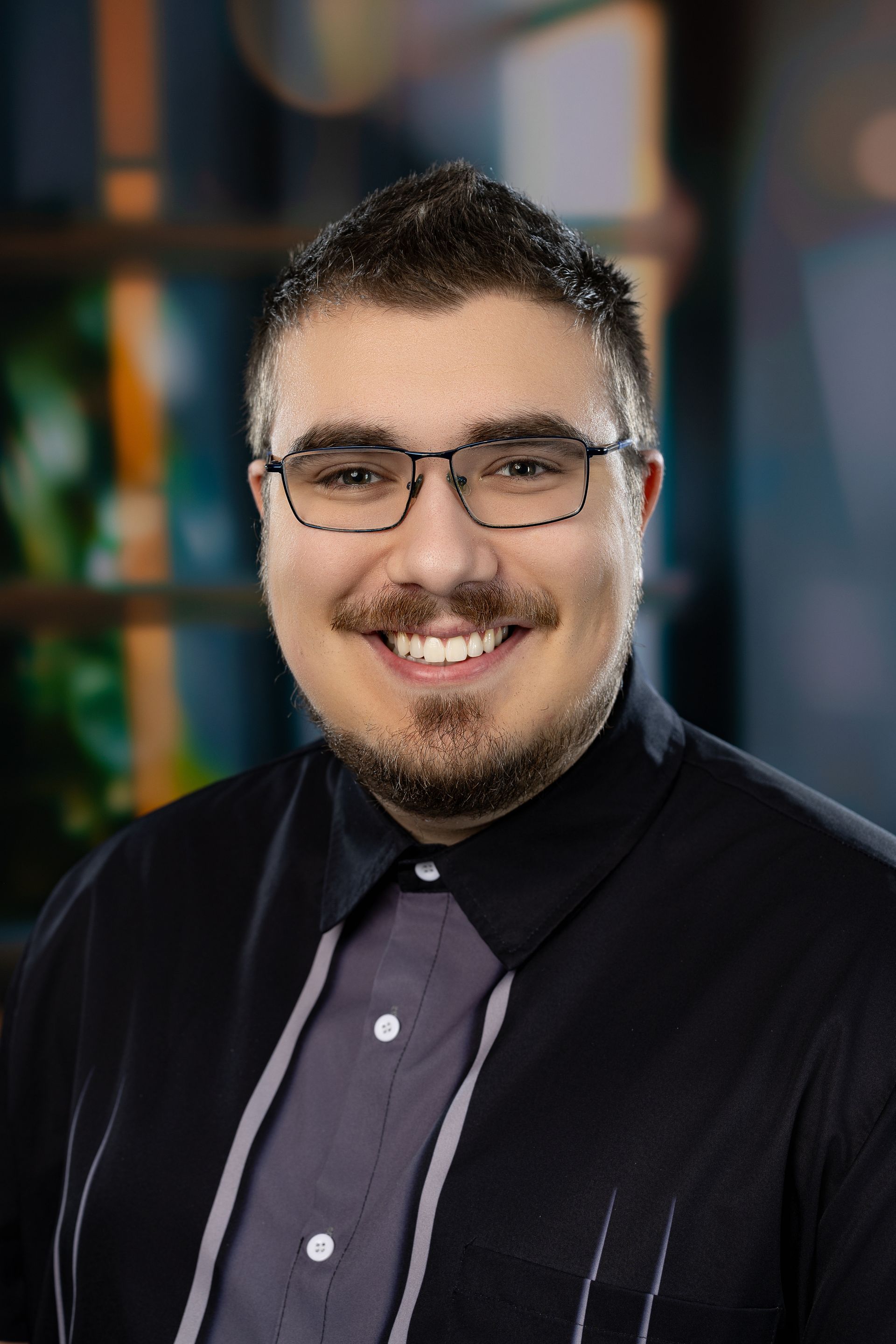 Un jeune homme portant des lunettes et une chemise bleue sourit à la caméra.