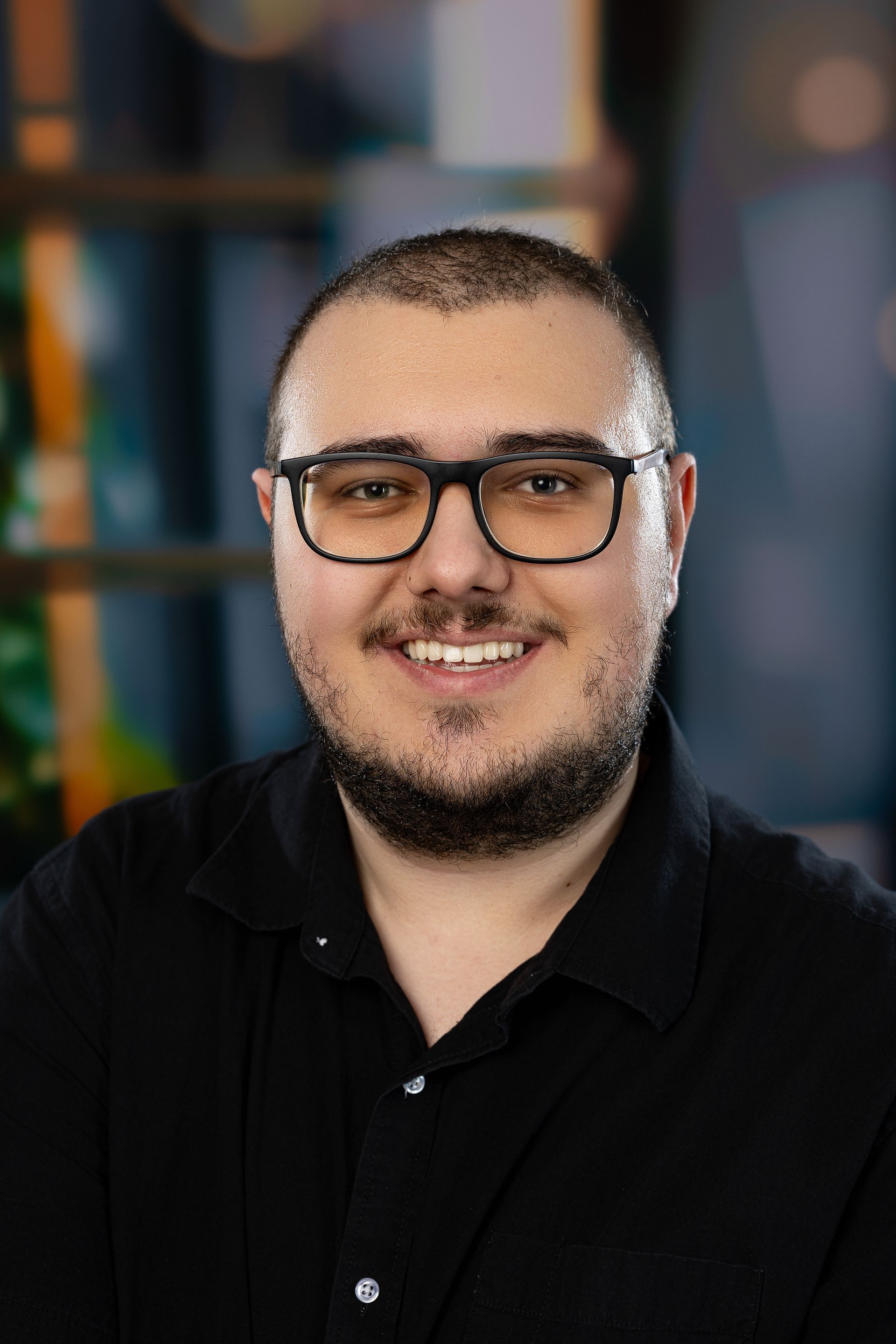 Un jeune homme avec des lunettes et une barbe sourit à la caméra.