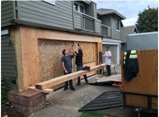 Men installing plywood over a garage door opening. Exterior shot of a house; gray siding, blue sky.