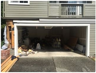 Exterior view of a damaged garage; exposed framing and debris, items stored inside, building siding above.