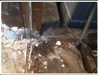 Damaged wooden floor with debris, pipes, and a partially visible fixture, suggesting a water leak in a dark, indoor space.