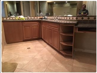Bathroom with light-brown cabinets, tan tile, and a dark countertop. A person is visible in the mirrors.