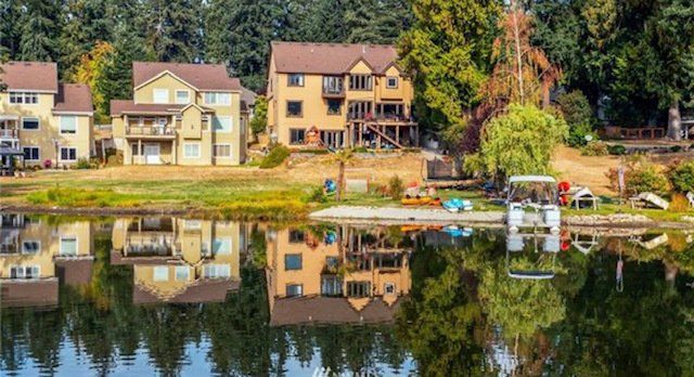 Lakeside houses with reflections in the water. Brown roofs, beige walls, green trees. Boat dock, sunny day.
