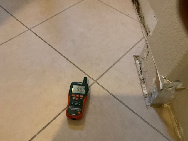 A moisture meter displaying 99 is on tiled floor next to damaged wall.
