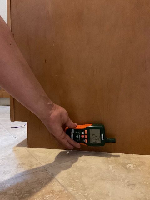 A person measures a wooden cabinet base with a black and orange level on a tile floor.