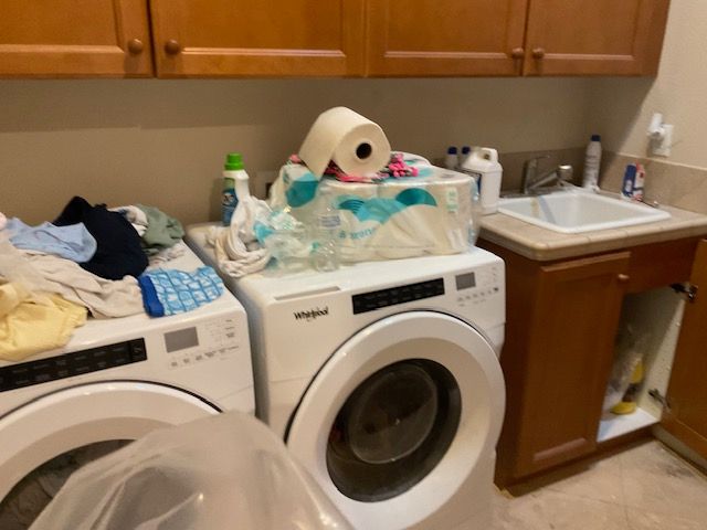 Laundry room with a washing machine and dryer, stacked with items including laundry, paper towels, and cleaning supplies.