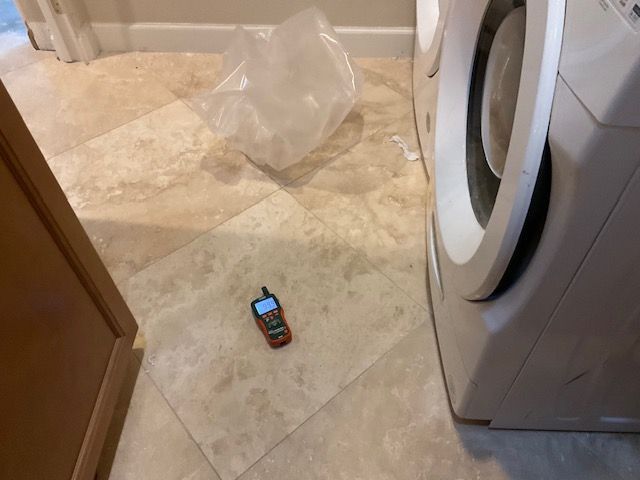 Cell phone on a tiled floor next to a washing machine, near a cabinet and a crumpled plastic bag.