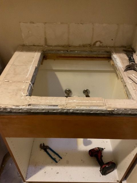 Bathroom vanity: tile countertop removed, cabinet below open, tools present.