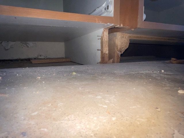 Underneath a kitchen cabinet, a wooden support post sits on a concrete floor.