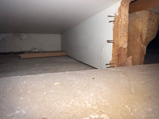 Crawlspace view showing a concrete floor, wooden beam with nails, and white wall.