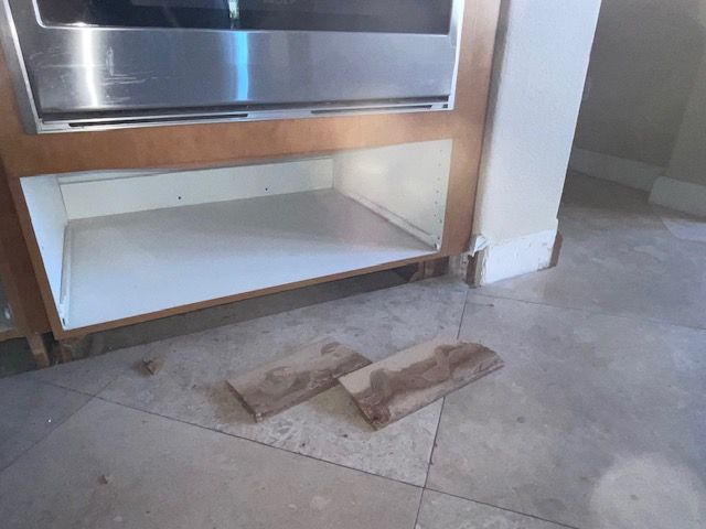 An open kitchen cabinet with two detached floor tiles on the floor in front of it.