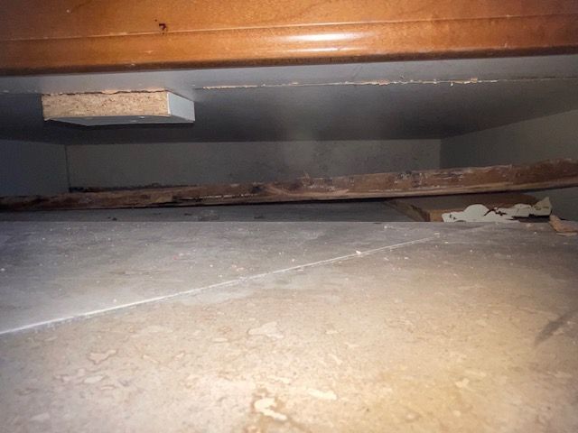 Under cabinet view: Partially visible brown cabinet above rotted wood and white tile floor.