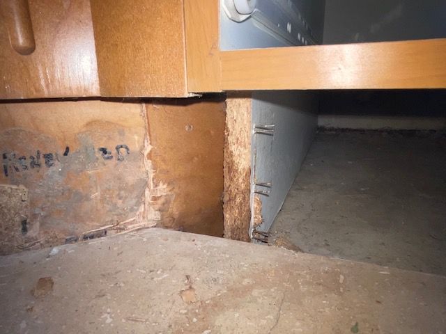Close-up of cabinet base showing damaged particle board and exposed floor.