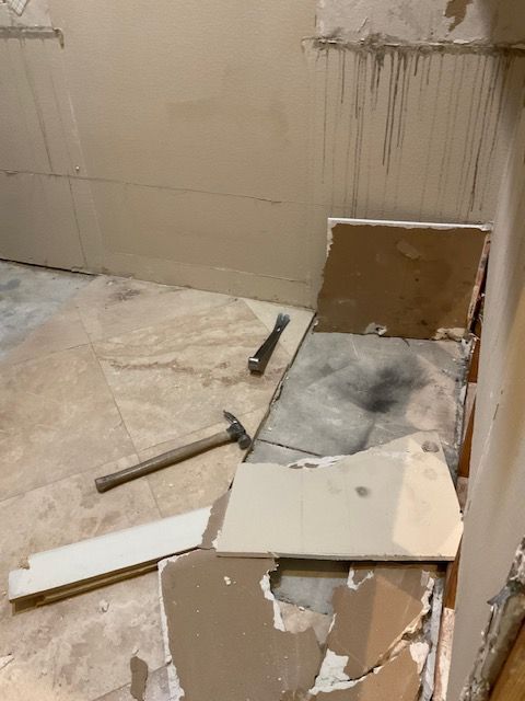 Room renovation in progress; damaged tile, debris, hammer and chisel on floor. Light beige walls.