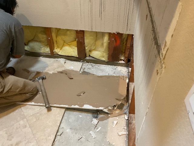 A person demolishes a bathroom floor. The wall is open, revealing insulation. Beige tiles and walls.