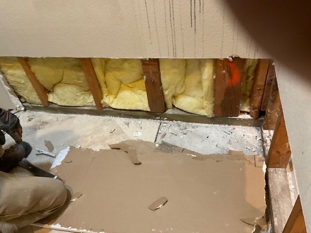Man removing drywall in a room, exposing wooden beams, insulation, and the floor.