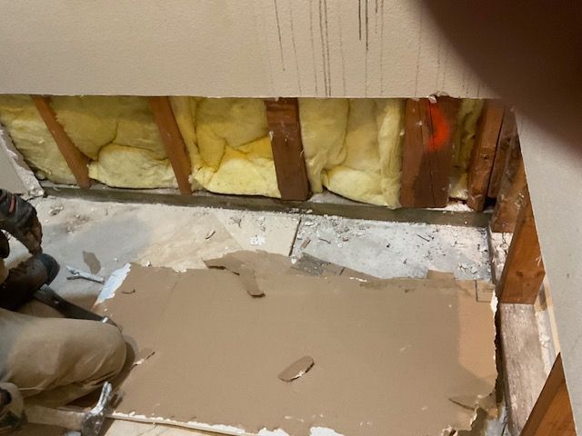 Person repairing a floor, removing material. Exposed insulation and wood beams visible.