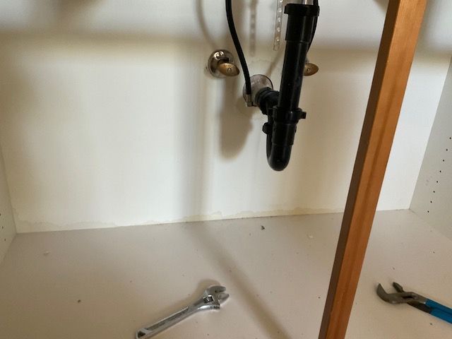 Inside of a white sink cabinet with plumbing, a wrench, and pliers.