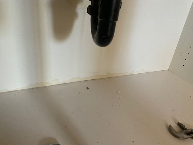 Under-sink cabinet with a black pipe and light-colored walls showing signs of water damage near the bottom.