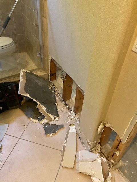 Bathroom wall damaged, exposing studs. Debris on tiled floor; shower and toilet visible.