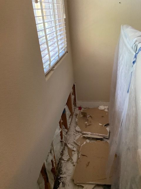 Damaged wall with exposed wood, debris, and a leaning mattress near a window.