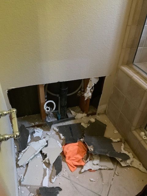 Bathroom wall ripped open, revealing plumbing. Debris and orange cloth on floor. Light and beige colors.