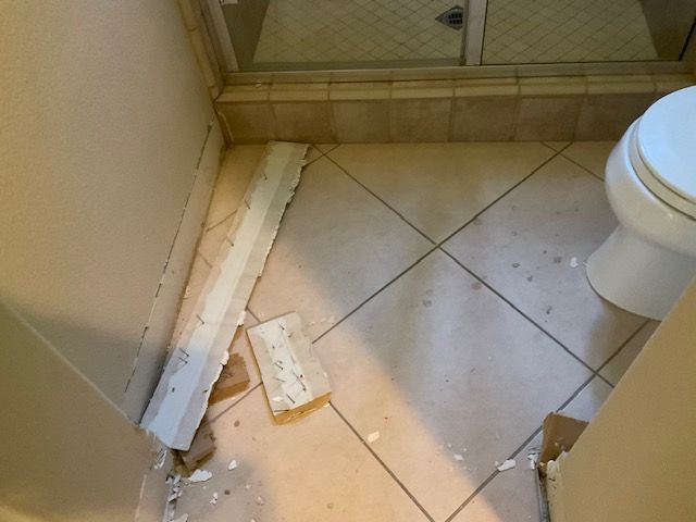 Bathroom floor with damaged baseboard trim, toilet on right, and shower in background.