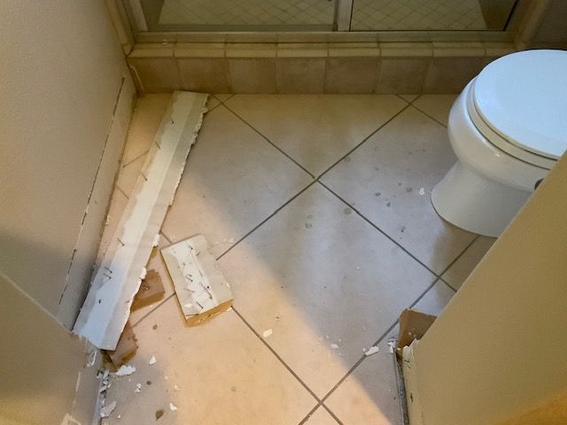 Bathroom floor with damaged baseboard and toilet. Beige tile, off-white walls.