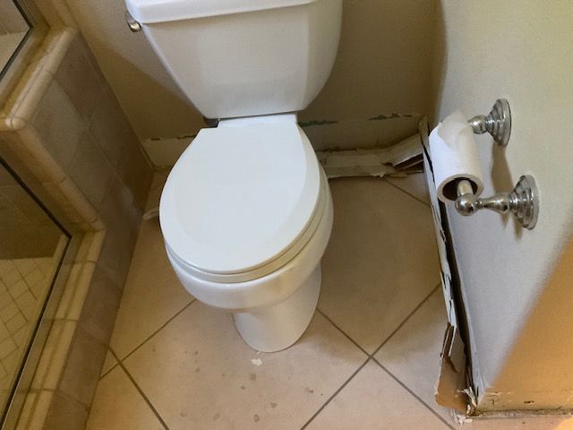 Toilet in a bathroom with damaged wall base and a mounted toilet paper holder.