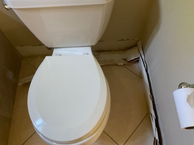 Toilet in a bathroom with peeling wallpaper, near a tile floor and toilet paper holder.
