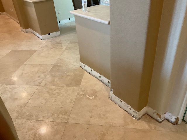 Beige tiled floor, walls, and countertops in a kitchen. Baseboards with holes.