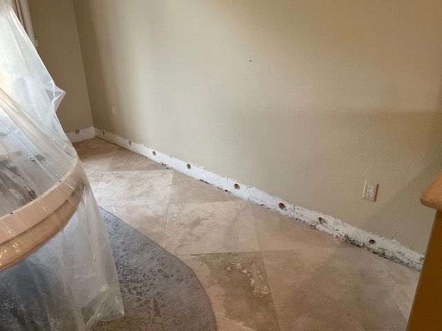 Room with tan and gray tiled floor, beige walls, and exposed baseboard with holes.